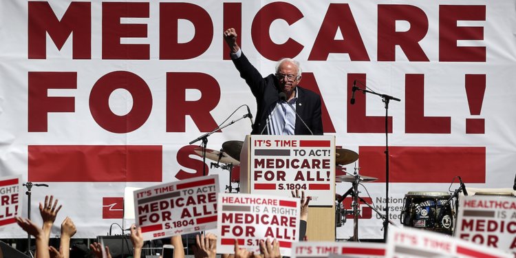 Bernie Sanders delivering a speech, surrounded by "Medicare for All" signs.