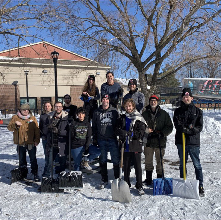 A mixed crowd of 13 people posing in a snowy field with several snow shovels.