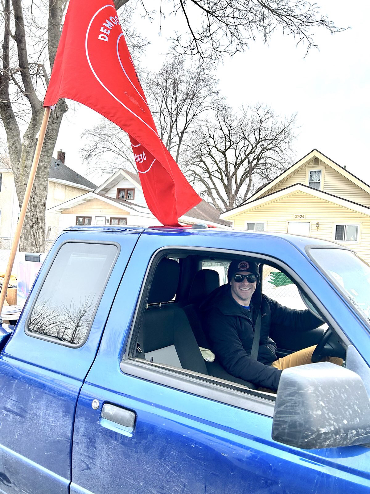 Smiling young white guy in blue truck with a red flag on it.